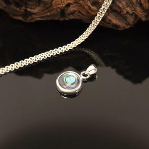 May include: A silver pendant necklace with a round, iridescent abalone shell center. The pendant is set in a polished silver bezel and hangs from a delicate silver chain. The background is dark, highlighting the necklace's details.