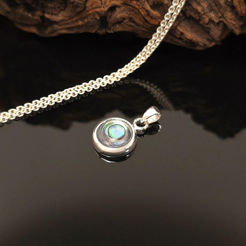 May include: A silver pendant necklace with a round, iridescent abalone shell center. The pendant is set in a polished silver bezel and hangs from a delicate silver chain. The background is dark, highlighting the necklace's details.