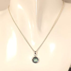 May include: A silver necklace with a small, round pendant. The pendant features a circular design with a light green and blue iridescent center. The chain is delicate and hangs on a white mannequin.