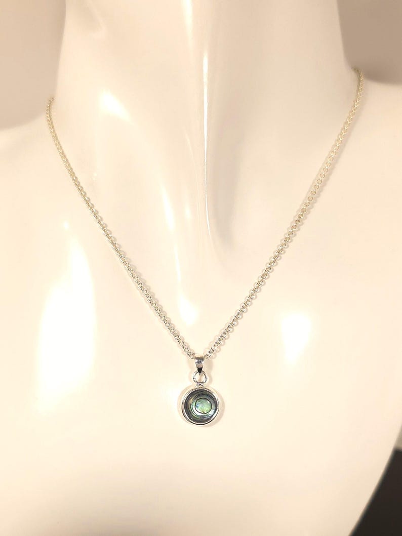 May include: A silver necklace with a small, round pendant. The pendant features a circular design with a light green and blue iridescent center. The chain is delicate and hangs on a white mannequin.