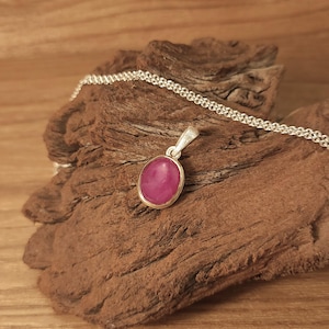 Peut inclure: Un pendentif en argent avec une pierre ovale rose. Le pendentif est posé sur une surface en bois.