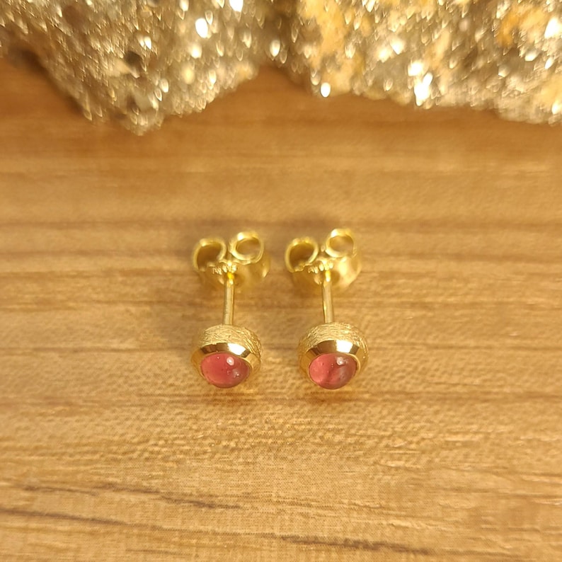 Puede incluir: Un par de pendientes de oro con piedras preciosas rosas. Los pendientes est&aacute;n sobre un fondo de madera.