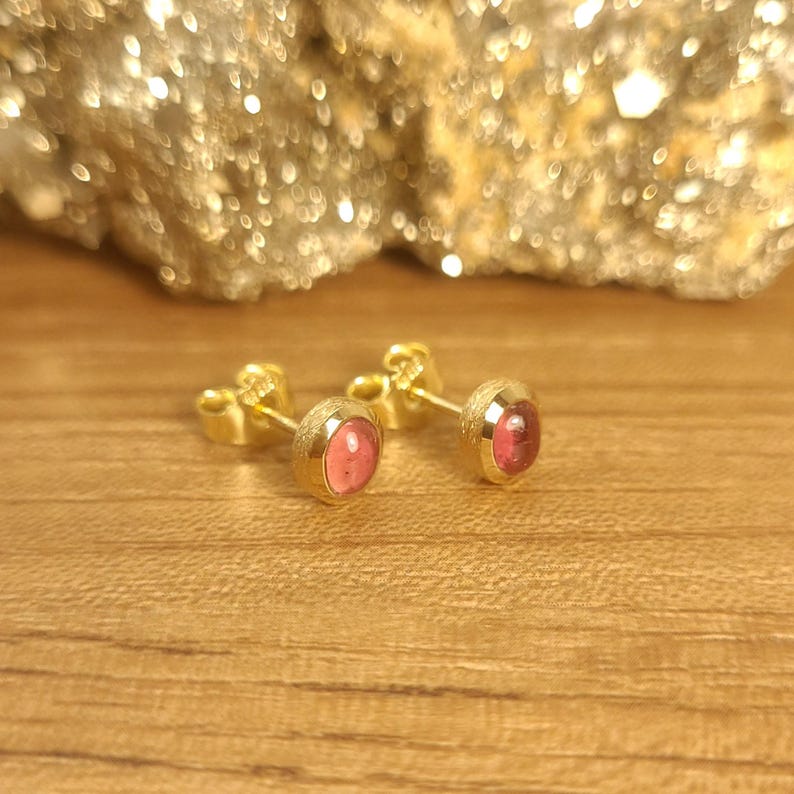 Puede incluir: Un par de pendientes de oro con piedras preciosas rosas. Los pendientes est&aacute;n sobre un fondo de madera.