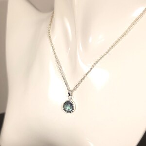 May include: A silver necklace with a small, round pendant. The pendant has a silver bezel and a shimmering, iridescent center, displaying shades of blue, green, and purple. The necklace is displayed on a white mannequin.