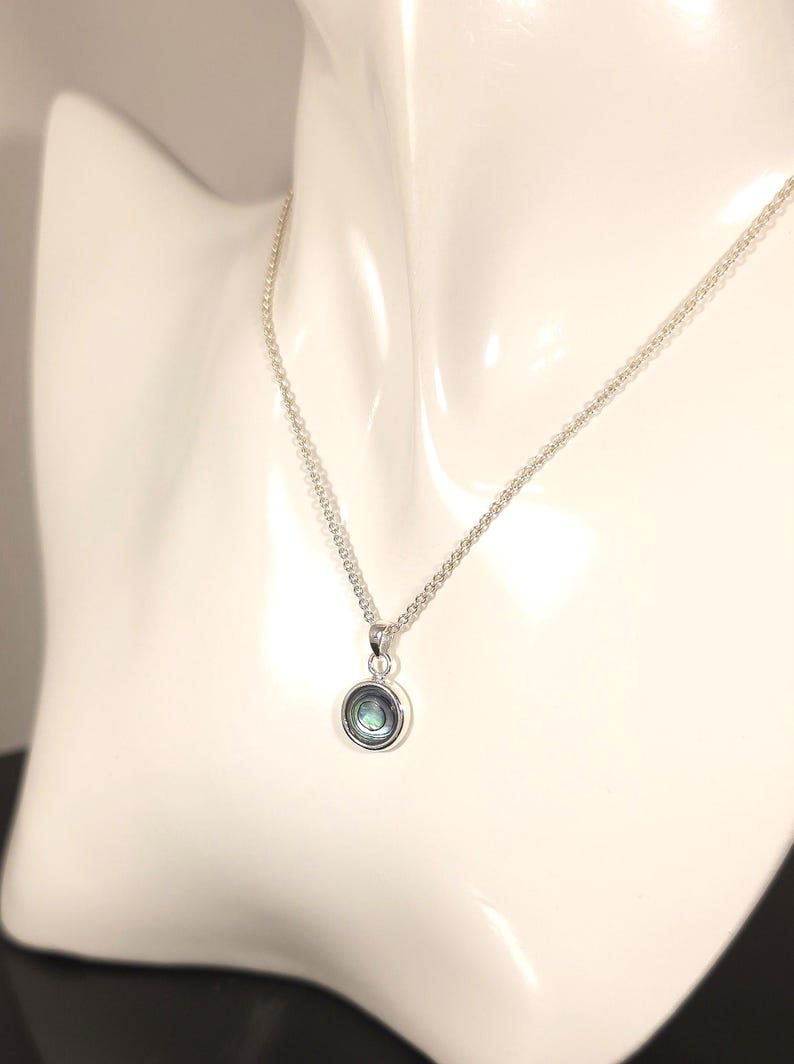 May include: A silver necklace with a small, round pendant. The pendant has a silver bezel and a shimmering, iridescent center, displaying shades of blue, green, and purple. The necklace is displayed on a white mannequin.