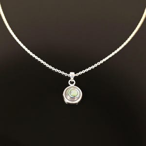 May include: A silver necklace with a small, round pendant. The pendant features a circular design with a pearlescent, iridescent center. The chain is delicate and the background is black.
