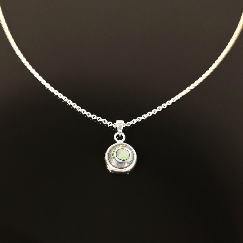May include: A silver necklace with a small, round pendant. The pendant features a circular design with a pearlescent, iridescent center. The chain is delicate and the background is black.