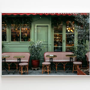 Könnte beinhalten: Foto eines Pariser Café-Außenbereichs. Das Café hat grüne Wände, eine rote Markise und Sitzgelegenheiten im Freien mit kleinen runden Tischen und gestreiften Bänken. Topfpflanzen und dekorative Lichter tragen zum Ambiente bei.