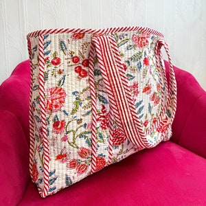 Op de afbeelding: Een witte gewatteerde tote bag met rood-wit gestreepte handvatten en een bloemenprint. De tas ligt op een roze fluwelen oppervlak.
