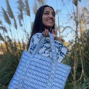 Puede incluir: Un bolso tote grande azul y blanco con un patrón de rayas. El bolso tiene dos asas y está hecho de una tela acolchada. La persona de la imagen lleva una chaqueta a juego con estampado azul y blanco.