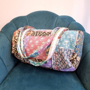 May include: A patchwork quilt duffel bag with a variety of colors and patterns, including pink, blue, green, and brown. The bag is sitting on a teal velvet chair.