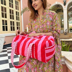 May include: A quilted duffel bag featuring alternating vertical stripes of bright red and light pink. The bag includes two matching striped fabric handles and a side pocket with a white and red checkered trim along the edges of the opening.
