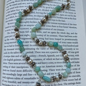 May include: A delicate necklace featuring pale blue-green gemstone beads, interspersed with pearls and silver-toned accents. The necklace is displayed on an open book, showcasing its elegant design and craftsmanship.
