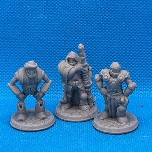 May include: Three gray miniature figurines, each standing on a round base. The figurine on the left is a robot with a helmet and a tool belt. The figurine in the center is a cloaked figure holding a staff. The figurine on the right is a knight in armor holding a sword.
