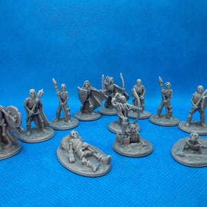 May include: A collection of gray miniature skeleton figurines in various poses, including standing, kneeling, and reclining. Some skeletons hold weapons like spears, swords, and bows, while others have shields. The figures are on circular bases against a blue background.