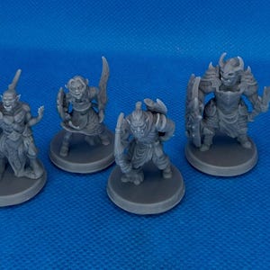 May include: Four gray miniature fantasy figures, likely for tabletop gaming. The figures include orc-like warriors with weapons and armor, standing on circular bases. The background is a solid blue.