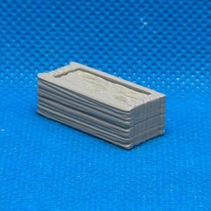 May include: A small, rectangular, gray object with a textured, layered appearance, resembling a miniature stack of wood. The top surface has a recessed rectangular area. The object is set against a solid blue background.
