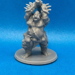 May include: A gray miniature figurine of a muscular orc-like creature, posed with arms raised. The figure has detailed armor with spikes and is standing on a gray circular base. The background is a solid blue.