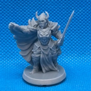 May include: A gray plastic miniature figurine of a warrior wearing armor and a cape, holding a sword. The figurine is standing on a round base.