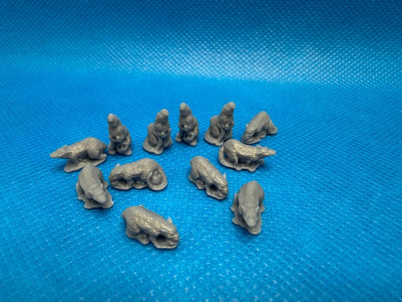 Op de afbeelding: Een verzameling van twaalf kleine, grijze, onbeschilderde plastic beeldjes die verschillende knaagdieren in diverse houdingen weergeven, waaronder enkele zittend en andere liggend, gerangschikt in een losse groep op een gestructureerd blauw oppervlak.