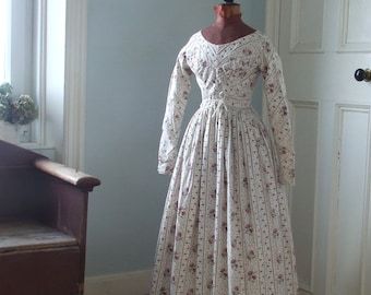 May include: A white dress with a floral print and vertical stripes. The dress has a fitted bodice and a full skirt. It is displayed on a mannequin in a room with a window and a door.