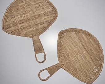 May include: Two hand-woven, natural-colored rattan fans. Each fan has a rounded, leaf-like shape with a woven pattern and a handle with a loop. The fans are displayed against a white background.