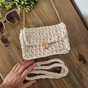 May include: A beige crocheted crossbody bag with a gold chain strap and a gold clasp. The bag has a long, beige crocheted strap that can be used as a shoulder strap.