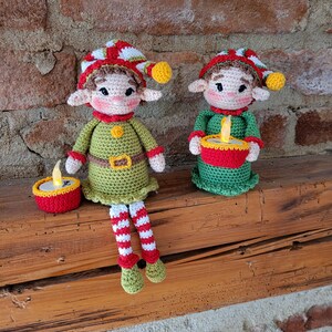 May include: Two crocheted elf dolls with festive hats and outfits. One elf sits with striped legs, the other holds a candle. Both have green and red accents, with yellow details. The dolls are handmade and perfect for holiday decor.