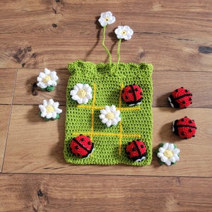 以下が含まれることがあります： 黄色の線と白いデイジーの花が付いた緑色の編み物製の三目並べゲームバッグ。黒い斑点と赤い体を持つ編み物製のテントウムシが6匹あり、そのうち4匹はバッグの上に、2匹はバッグから離れています。