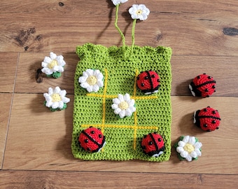 May include: A green crocheted tic-tac-toe game bag with yellow lines and white daisy flowers. There are six crocheted ladybugs with black spots and red bodies, four of which are on the bag and two are off the bag.