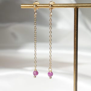 Peut inclure: Boucles d'oreilles pendantes en chaîne dorée avec de petites pierres précieuses roses et rondes en bas. Les boucles d'oreilles sont suspendues à une barre dorée. Le fond est un tissu blanc et doux.