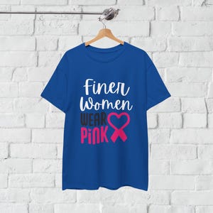 May include: A royal blue t-shirt with the text "Finer Women Wear Pink" in white and pink. The design includes a pink ribbon and heart symbol, displayed on a wooden hanger against a white brick wall.