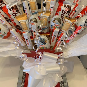 May include: A bouquet of Kinder chocolate treats arranged in a red box with a white bow. The bouquet includes Kinder Surprise eggs, Kinder Bueno bars, Kinder Happy Hippo bars, and other Kinder chocolate products.