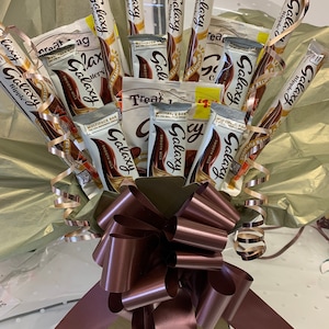 May include: A bouquet of chocolate bars wrapped in gold and brown paper with a brown satin bow. The chocolate bars are all Galaxy brand and feature the text "Galaxy" and "Smooth Milk".