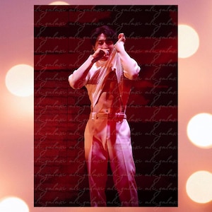 May include: A man wearing a white shirt and white pants poses for a photo. He is wearing a white shirt that is partially pulled off his shoulders, revealing his toned torso. The background is a red and white pattern.