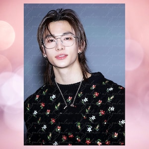 May include: A portrait of a person wearing eyeglasses, a black sweater with floral embroidery, and a silver necklace. The person has long, dark hair and is looking at the camera. The background is a muted gray with a watermark.
