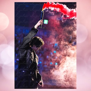 May include: A person holding a black flare emitting red smoke against a backdrop of blue and purple stage lighting. The person is wearing a dark jacket. The image has a watermark in the background.