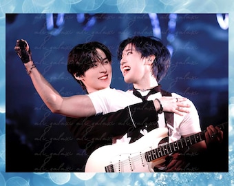 May include: Two individuals on stage, one with dark hair wearing a white t-shirt and black jacket, the other with blue-tinted hair in a white t-shirt, embracing while one plays a white electric guitar. The scene is set against a dark blue background.