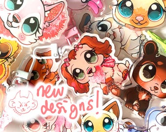May include: A collection of colorful cartoon animal keychains with large eyes. The keychains are made of clear acrylic and feature different animals, including a dog, a cat, a bear, and a fox. The text "new designs!" is written in pink on a white background.