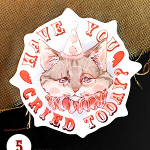 K&ouml;nnte beinhalten: Ein wei&szlig;er runder Aufkleber mit rotem Rand und dem Text "Hast du heute geweint?" in einer roten, stilisierten Schrift. Eine Comic-Katze mit einer Partym&uuml;tze und einem roten und wei&szlig;en Kragen befindet sich in der Mitte des Aufklebers. Die Katze hat einen traurigen Ausdruck und schaut nach unten.