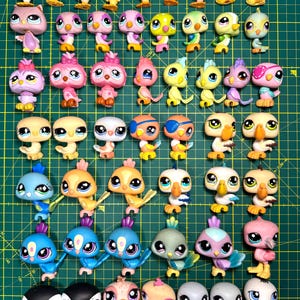 Puede incluir: Una colección de pequeñas figuras de pájaros de colores, con grandes ojos. Los pájaros son de varios colores, incluyendo amarillo, rosa, azul y naranja. Están dispuestos sobre una esterilla de corte verde con marcas en centímetros.