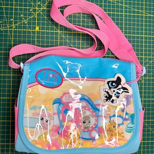 May include: A light blue and pink messenger bag featuring a clear front panel with a colourful Littlest Pet Shop graphic print. The bag includes a pink adjustable shoulder strap, a red oval LPS logo, and a dangling white dog-shaped tag.