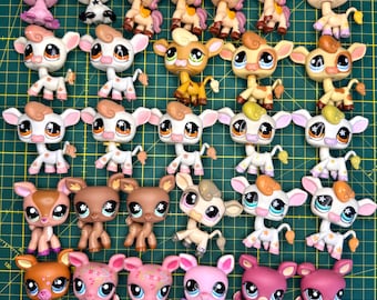 May include: A selection of small plastic animal figurines in various colours, including pigs, ponies, cows, and deer. The toys are in shades of pink, brown, and white, with large eyes. The figurines are arranged on a green grid surface.