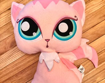May include: A plush toy shaped like a cat with light pink fur, large teal and black eyes, and a small pink nose. The cat has pointed ears, a white chest patch, and a striped tail. A small white tag with black text is attached to the side.