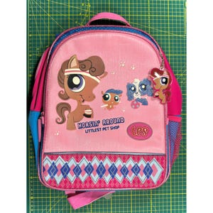 以下が含まれることがあります： 漫画の馬と犬のグラフィックが特徴のピンク色のバックパック。テキストは「Horsin' Around Littlest Pet Shop」です。バックパックには青とピンクのアクセント、上部のハンドル、ひし形模様のフロントポケットが付いています。