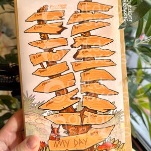 Op de afbeelding: Een notitieboek met een handgetekende illustratie van een boom met houten wegwijzers die tijden van 5 uur tot 22 uur aangeven. De woorden "MY DAY" staan onderaan, met een eekhoorn en paddenstoelen. Het notitieboek wordt door een hand vastgehouden.