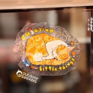 Puede incluir: Una pegatina transparente con un gato naranja y blanco durmiendo. El texto "enjoying little things" está escrito en amarillo. El logotipo de Outdoor Seekers está en la parte inferior.