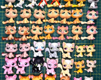 May include: A collection of small plastic cat figurines with large eyes, arranged in rows on a green cutting mat. The figures feature various colors including pink, orange, white, gray, and black, with some displaying distinct patterns and markings.
