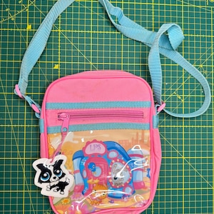 Könnte beinhalten: Eine pinke Umhängetasche mit hellblauem, verstellbarem Riemen und Besatz. Die Vorderseite hat eine durchsichtige Kunststofftasche mit einer bunten Littlest Pet Shop-Grafik und dem Text LPS. Ein kleiner, ausgestanzter Pappanhänger eines schwarz-weißen Hundes ist befestigt.