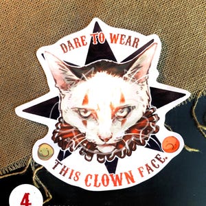 K&ouml;nnte beinhalten: Eine wei&szlig;e Katze mit orangefarbenen und schwarzen Markierungen, die ein rot-schwarzes Clownshalsband tr&auml;gt. Die Katze schaut den Betrachter mit einem ernsten Ausdruck direkt an. Das Bild ist ein Aufkleber mit dem Text "Dare to wear this clown face." und einem schwarzen Stern hinter der Katze. Der Aufkleber ist 7,2 cm x 7,2 cm gro&szlig;.